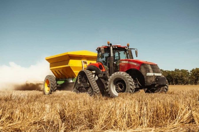 Un puissant tracteur agricole rouge à chenilles tracte une remorque d'épandage jaune dans un champ de chaume avant l'usage d'un tracteur enjambeur viticole.