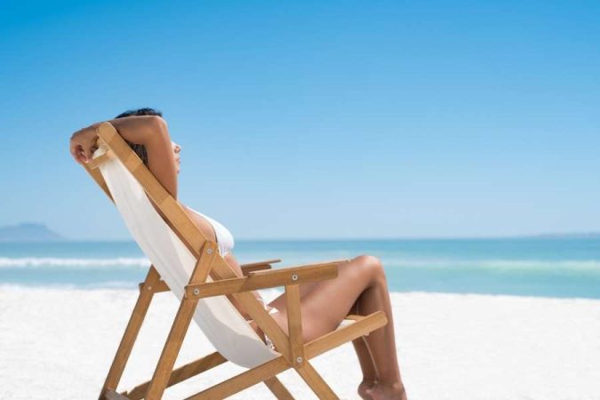 Une femme en maillot de bain blanc assise dans une chaise longue en bois sur une plage de sable blanc, face à la mer bleue turquoise, pour parfaire son bronzage.