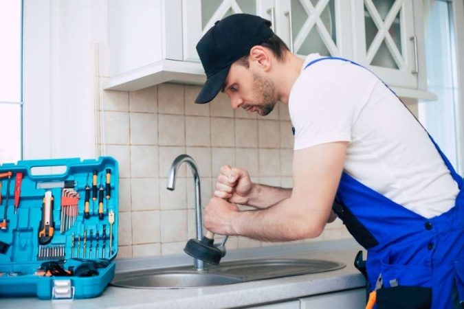 Un plombier casquette et combinaison bleue utilise une ventouse pour déboucher un évier de cuisine.