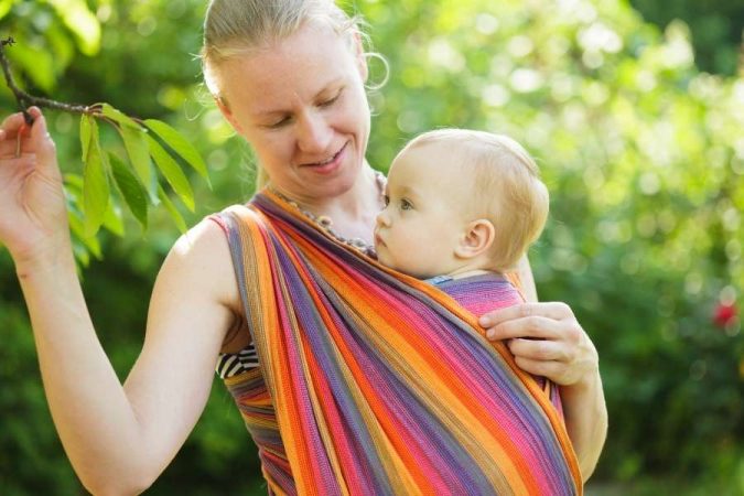 Une maman souriante pratique le portage physiologique avec son bébé dans une écharpe colorée en plein air.