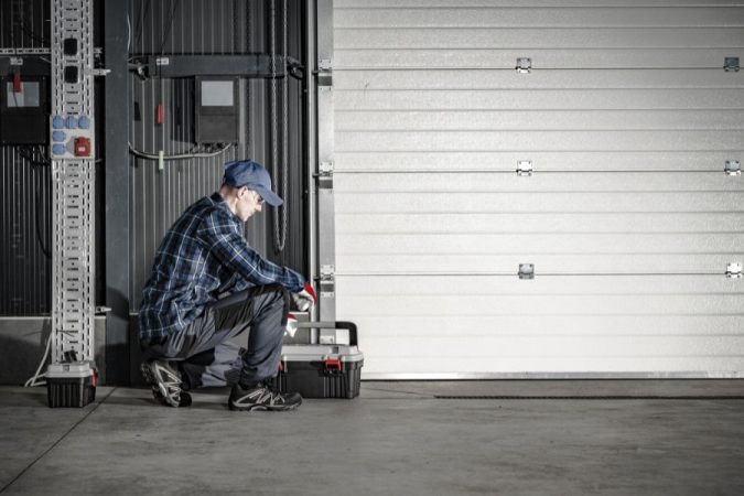 Un technicien accroupi avec sa boîte à outils s'apprêtant à sécuriser une porte de service ou de garage sectionnelle blanche.